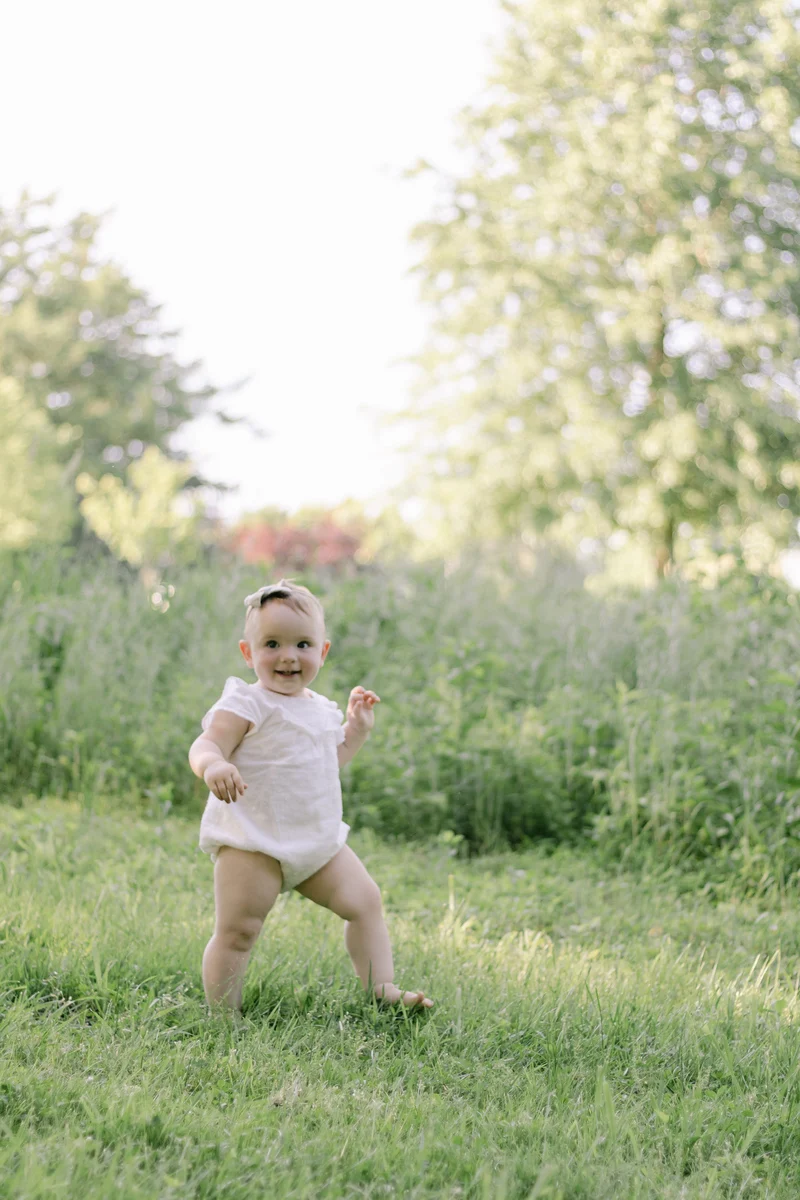 Smiling baby girl in a white romper taking her first steps across a grassy meadow in Bucks County