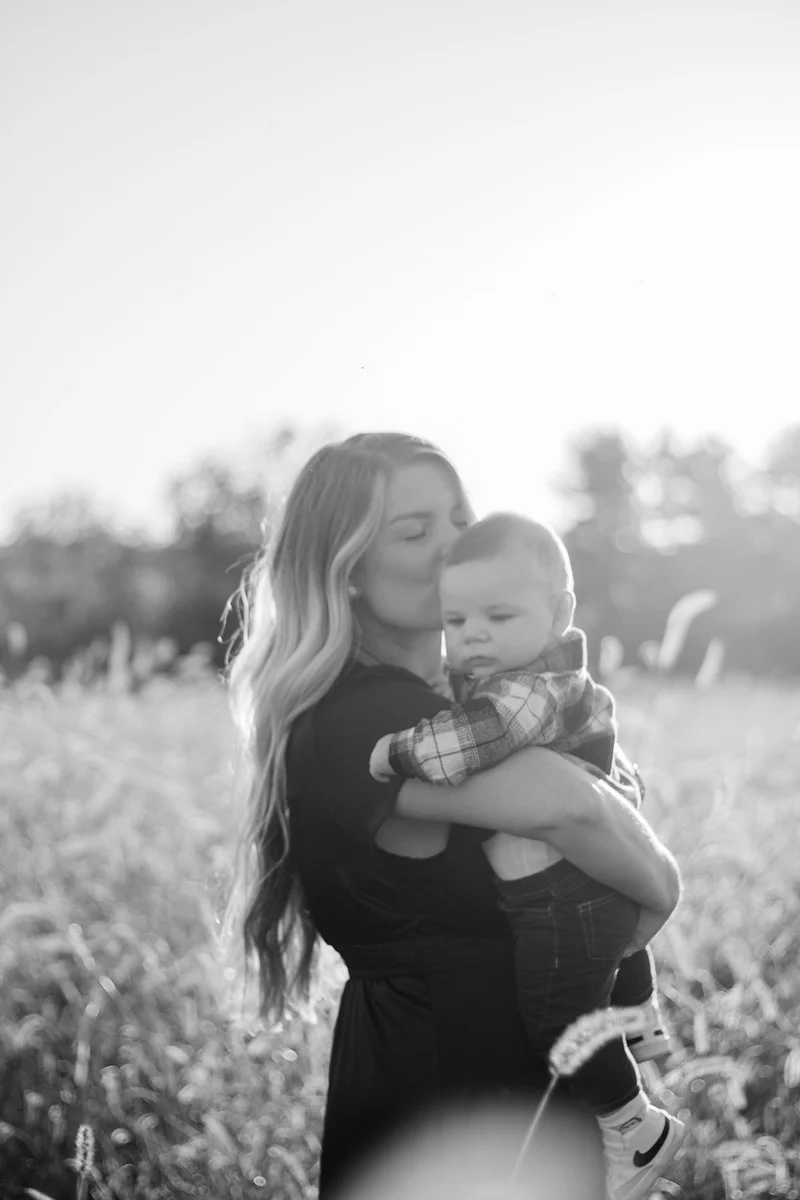 Black-and-white photo of a mother kissing her baby boy's head in a sunlit field