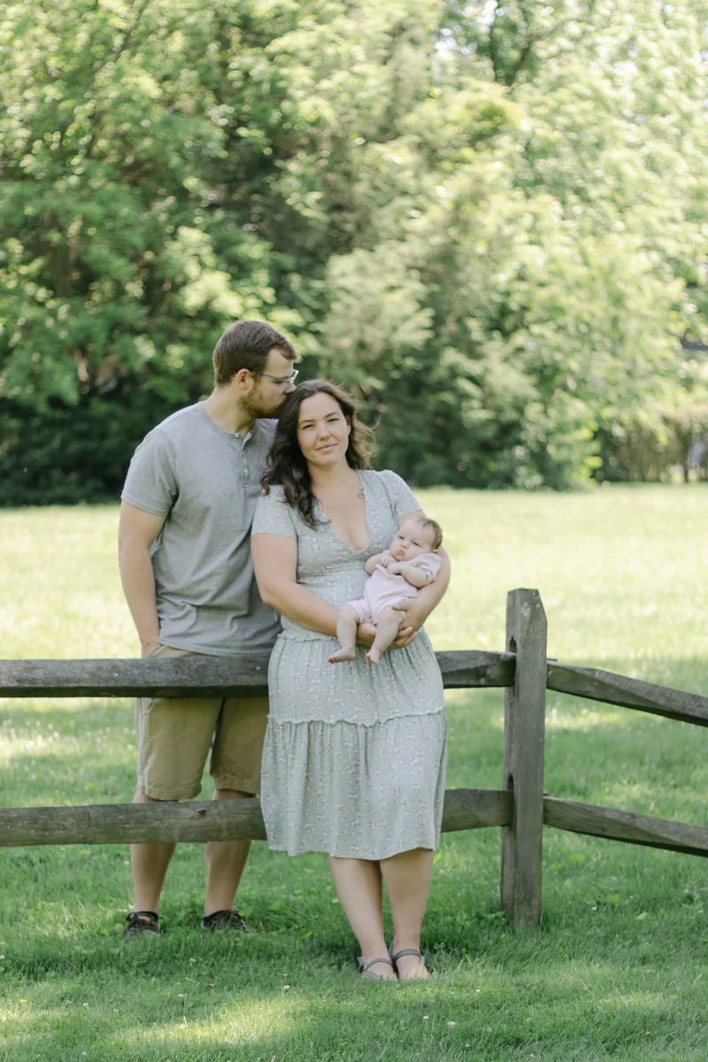 Mom and dad holding their newborn daughter beside a split-rail fence in a green Bucks County park