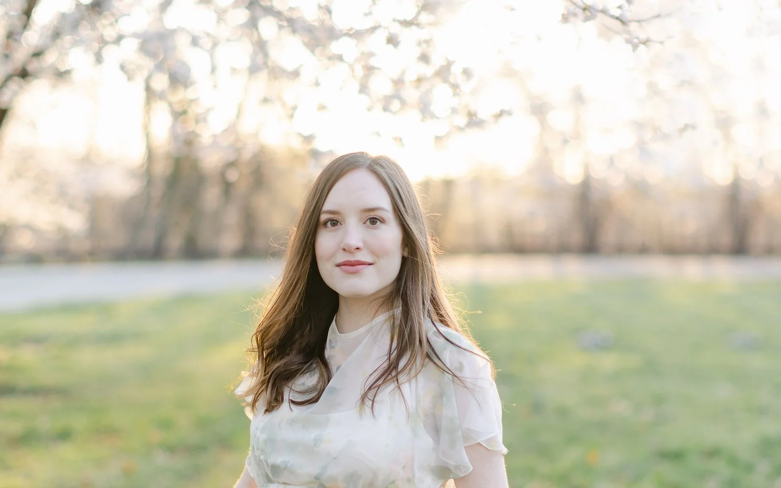 Woman in a soft floral dress smiling beneath blooming cherry blossoms at sunset