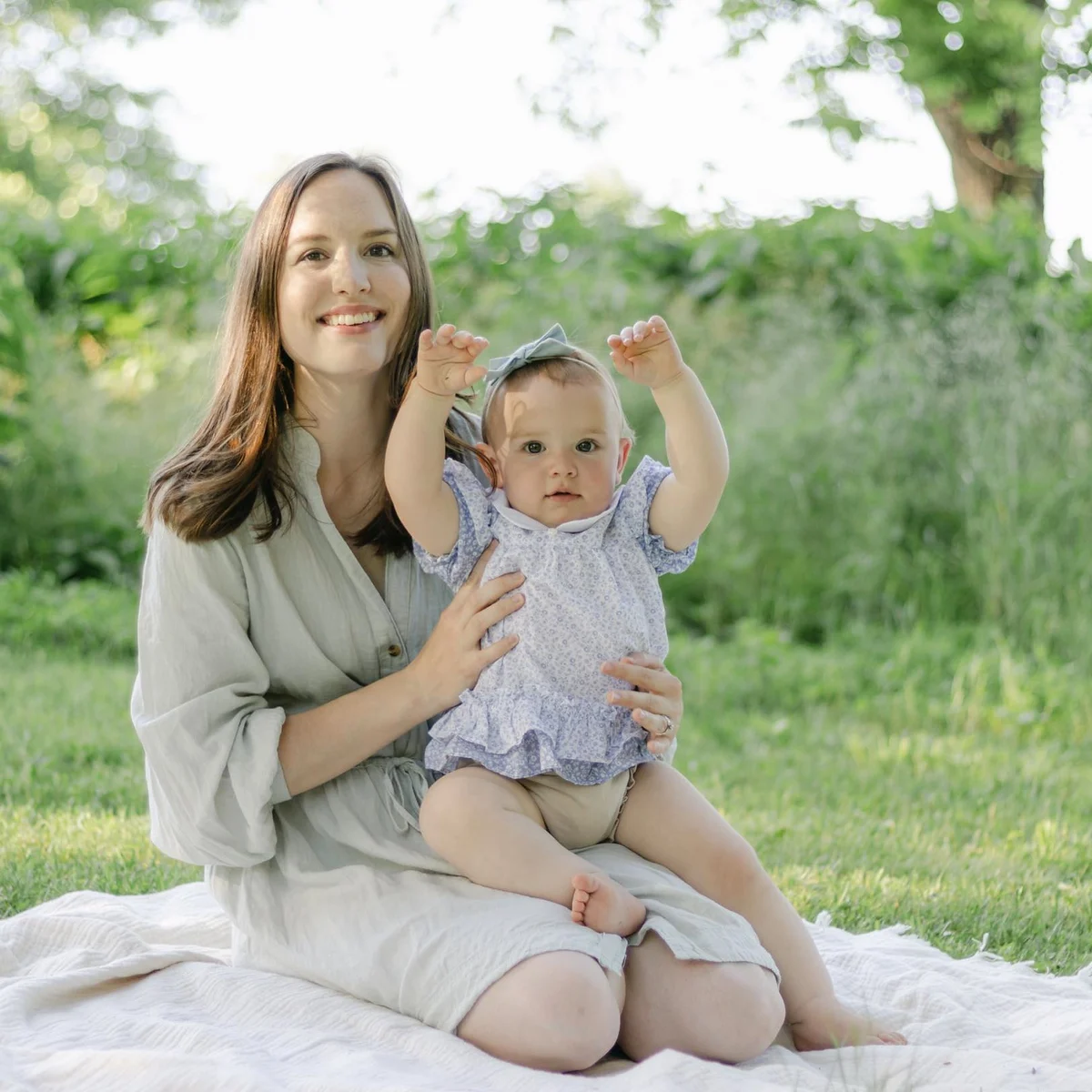 Mother sitting on a picnic blanket in the grass holding up her smiling baby daughter