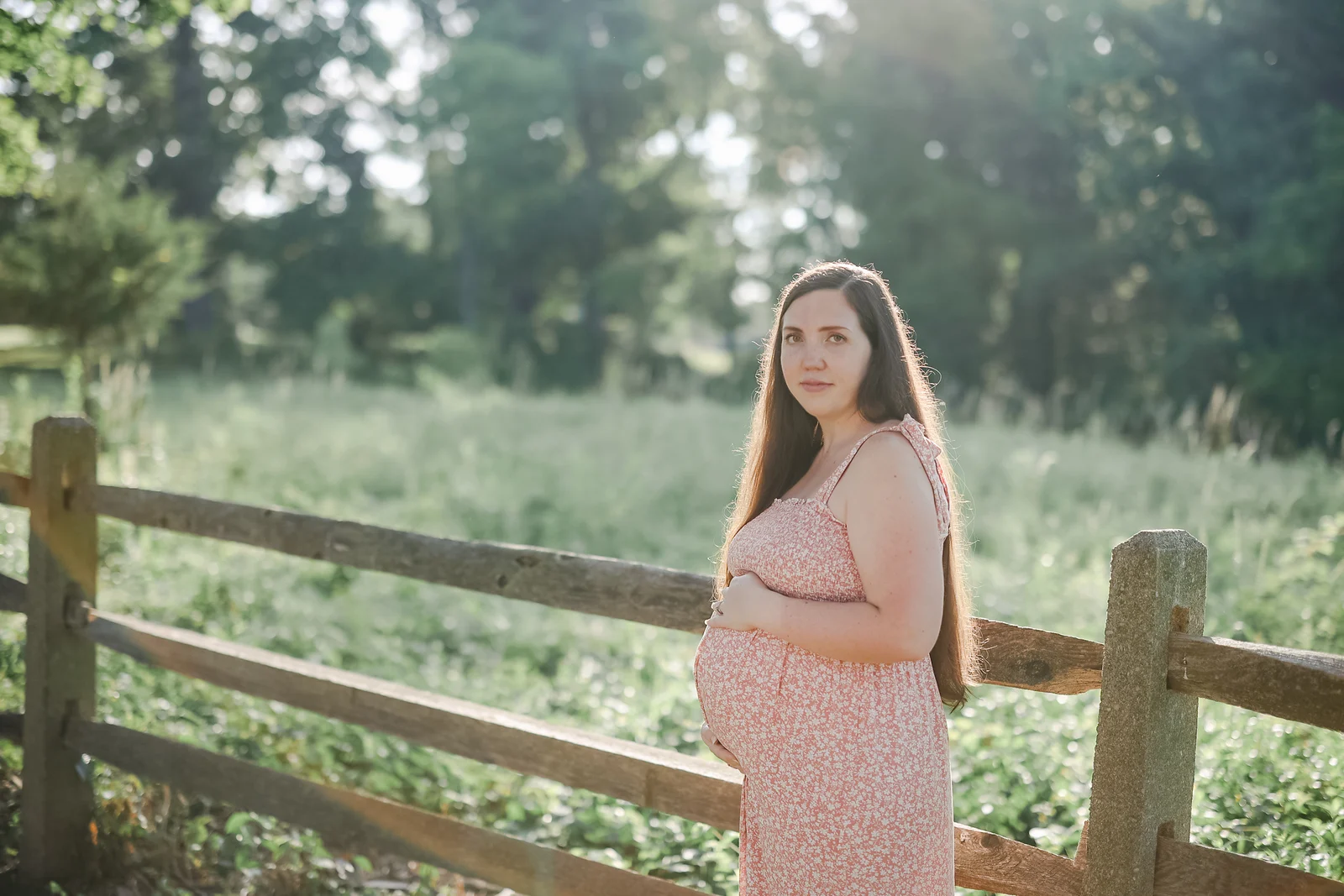 Expectant mother in a pink floral dress cradling her bump beside a split-rail fence in a summer meadow