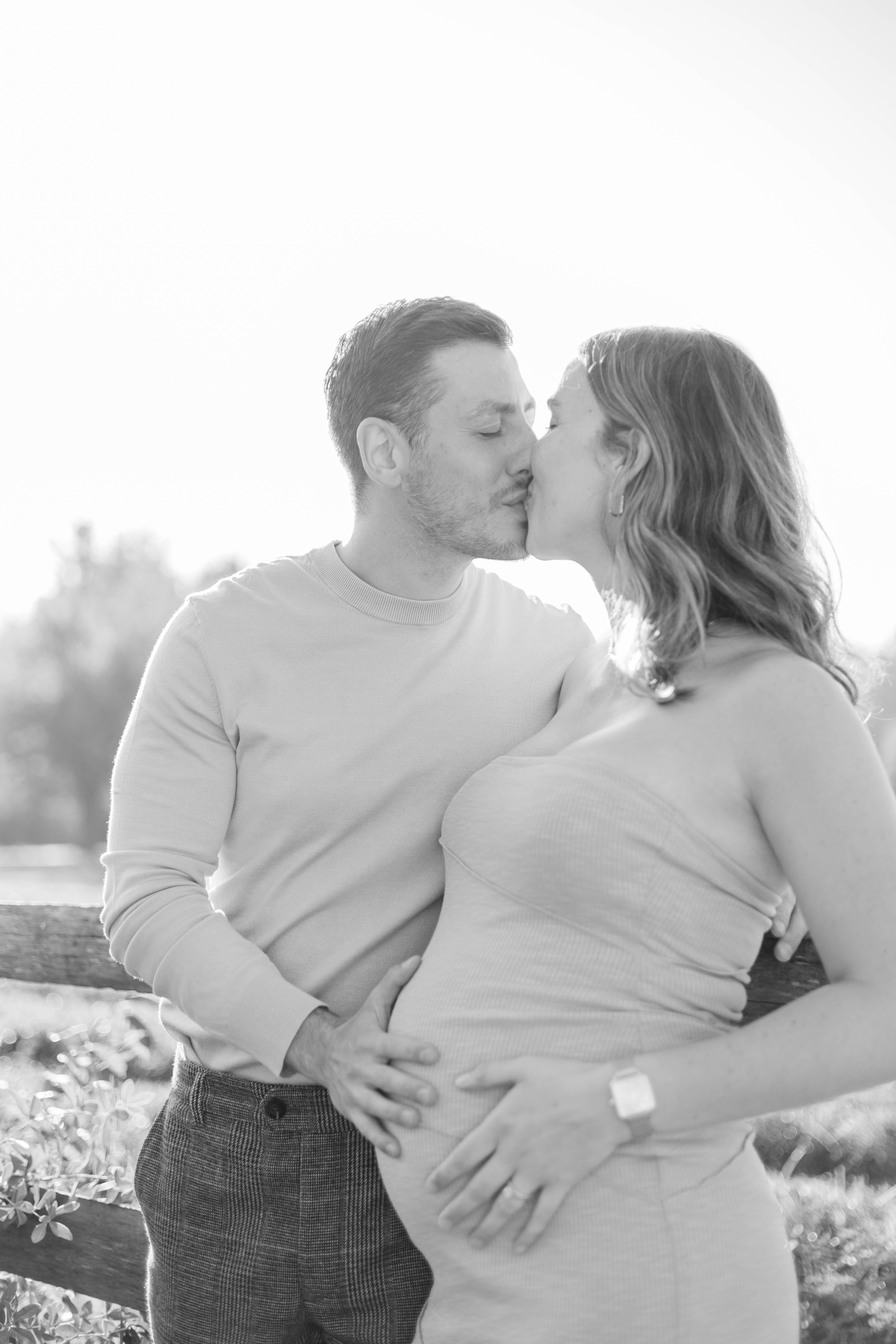 Black-and-white maternity portrait of an expectant couple kissing, both their hands resting on her bump