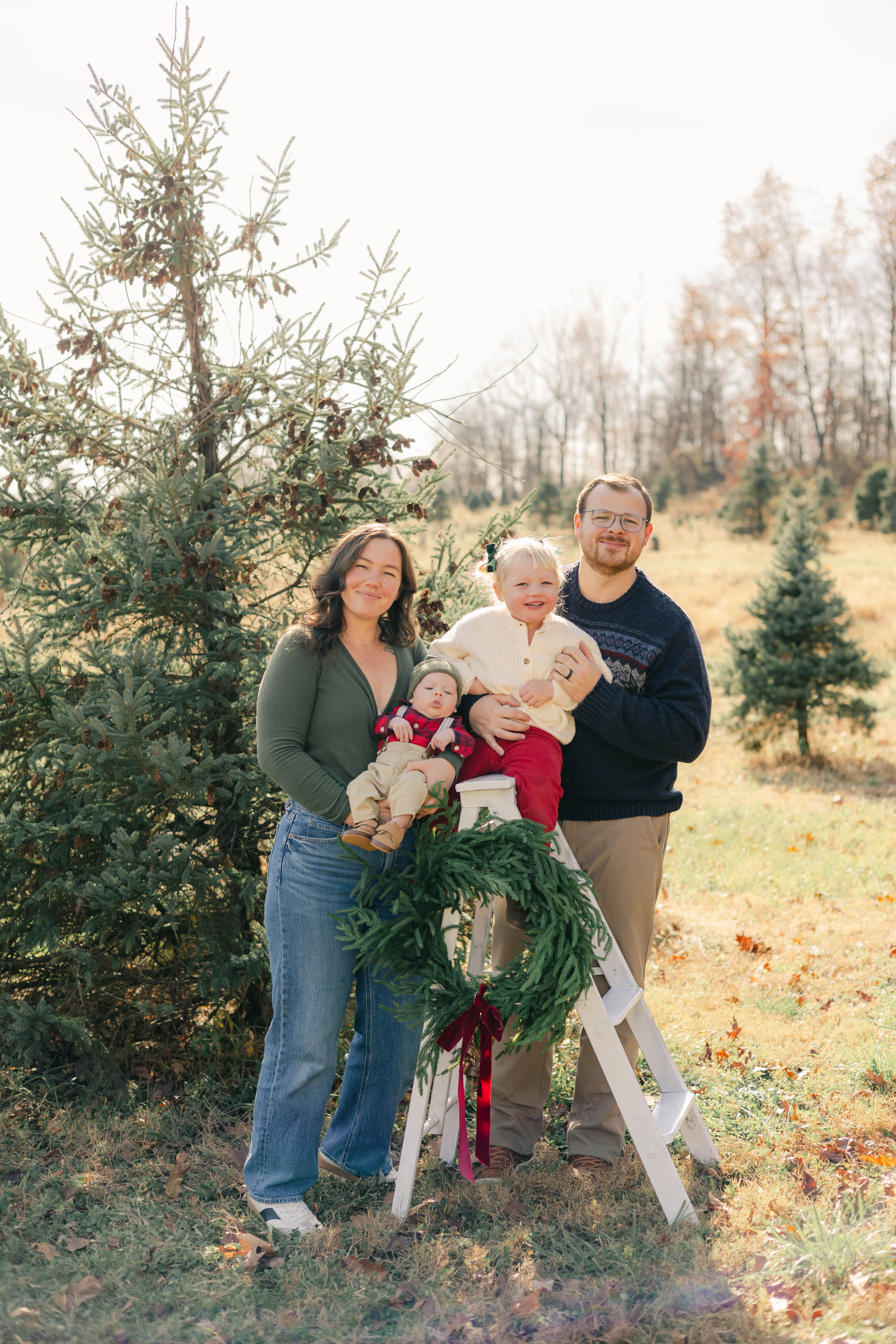 Family with two young children posing beside a wreath-decorated ladder at a Christmas tree farm