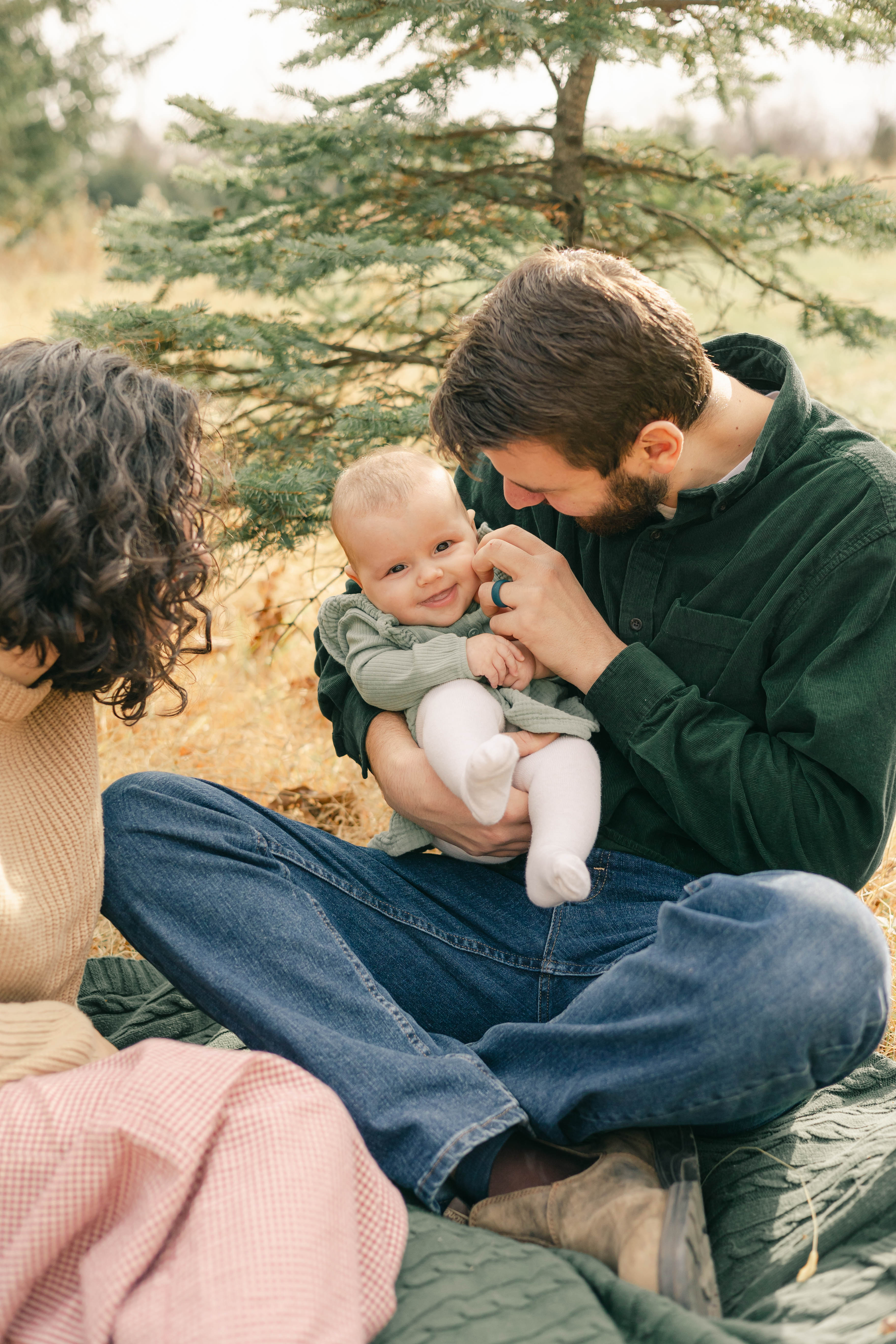 Dad cuddling his smiling baby on a picnic blanket beneath evergreens while mom looks on
