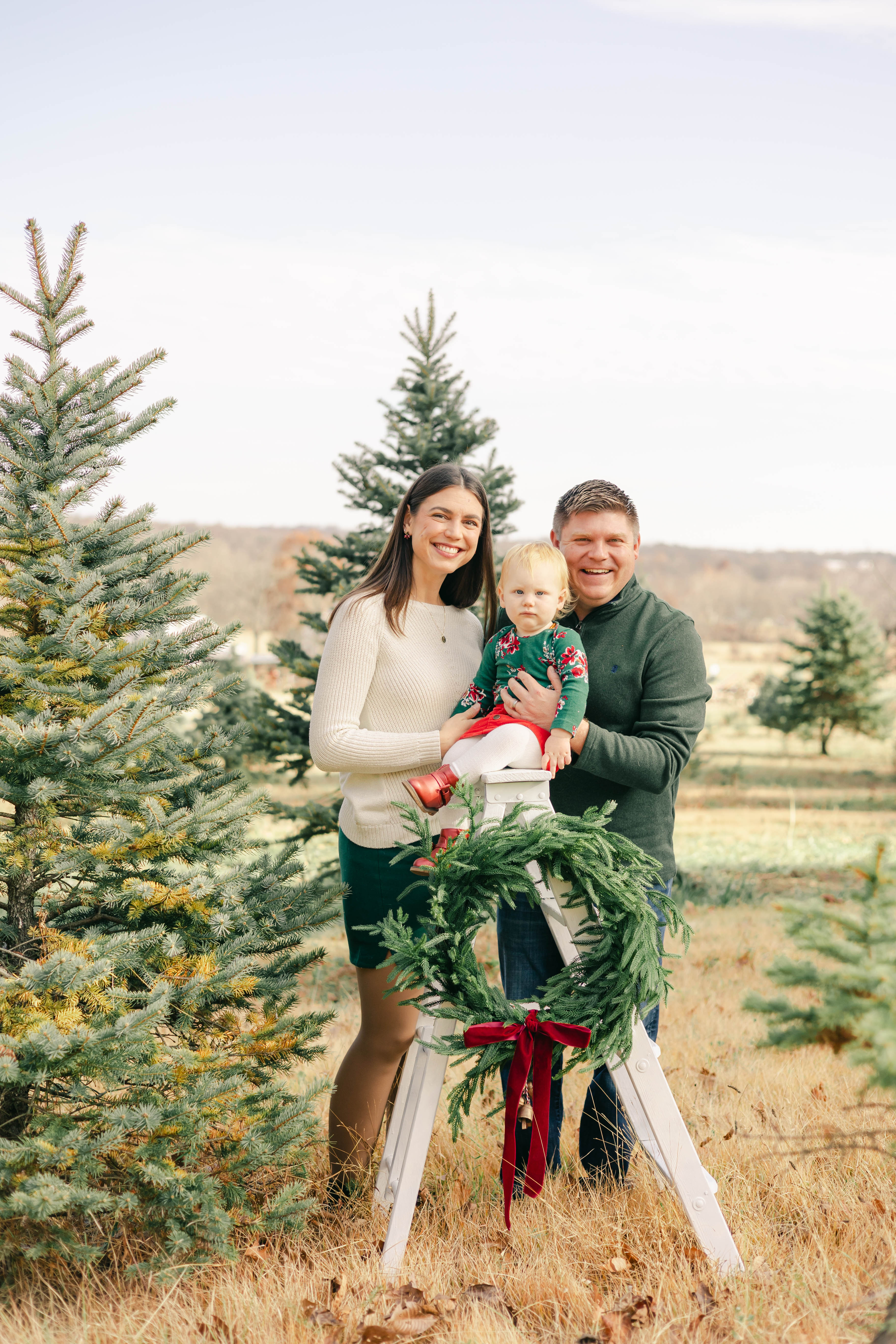 Parents holding their baby beside a wreath-trimmed ladder among Christmas trees