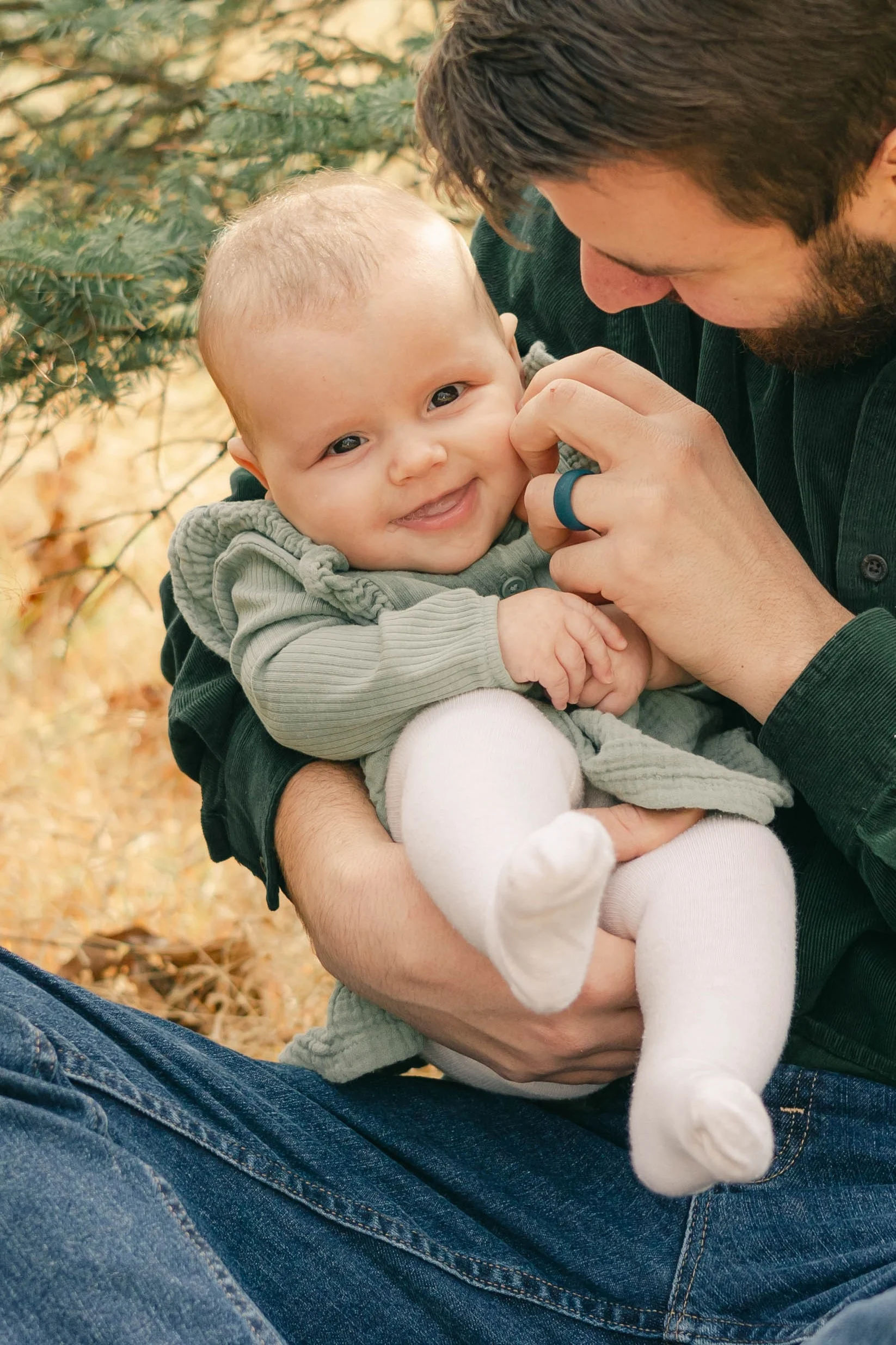 Smiling baby in a green knit sweater held on her dad's lap during an autumn outdoor session