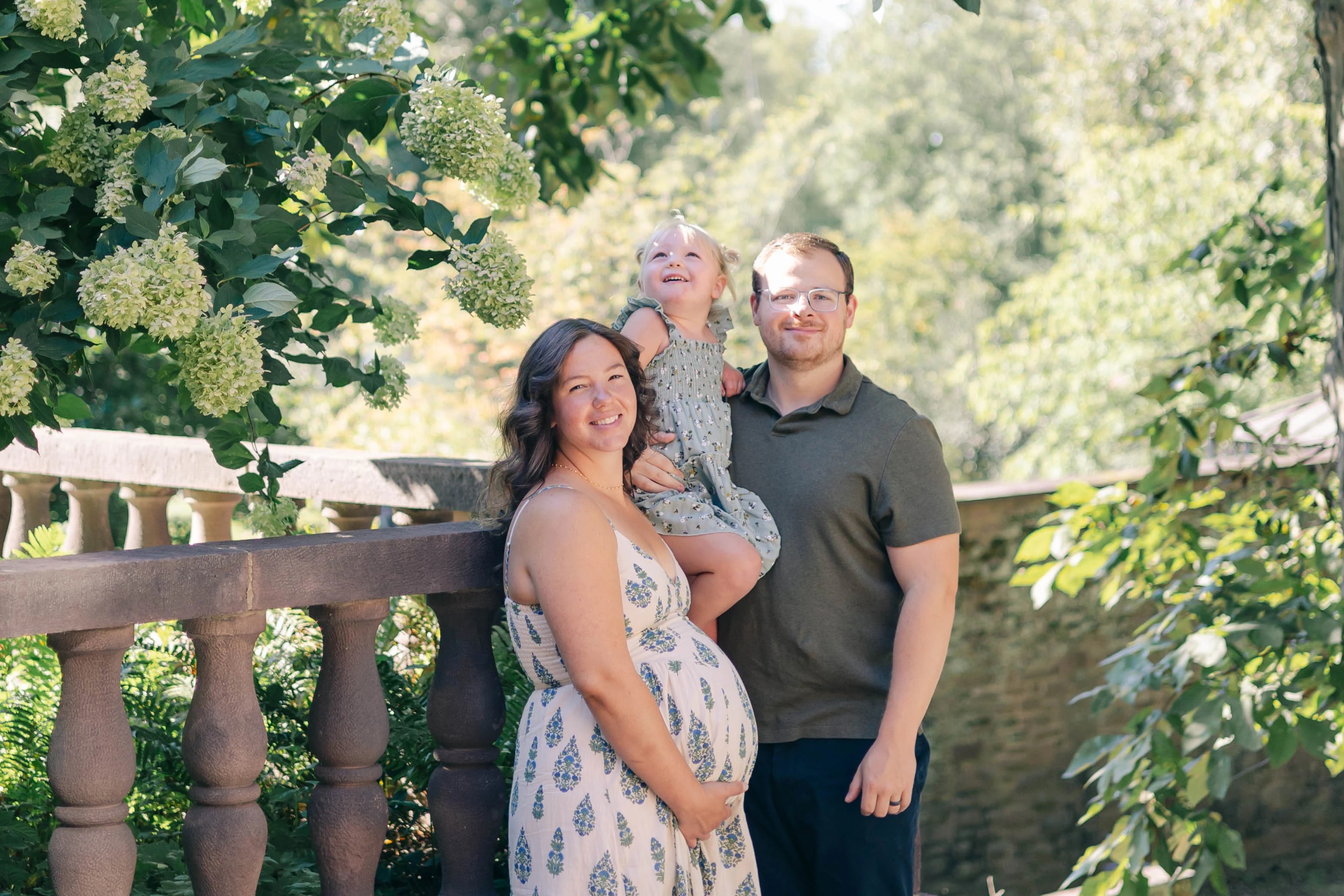 Expectant mom, dad and their toddler daughter beside a stone balustrade framed by white hydrangeas